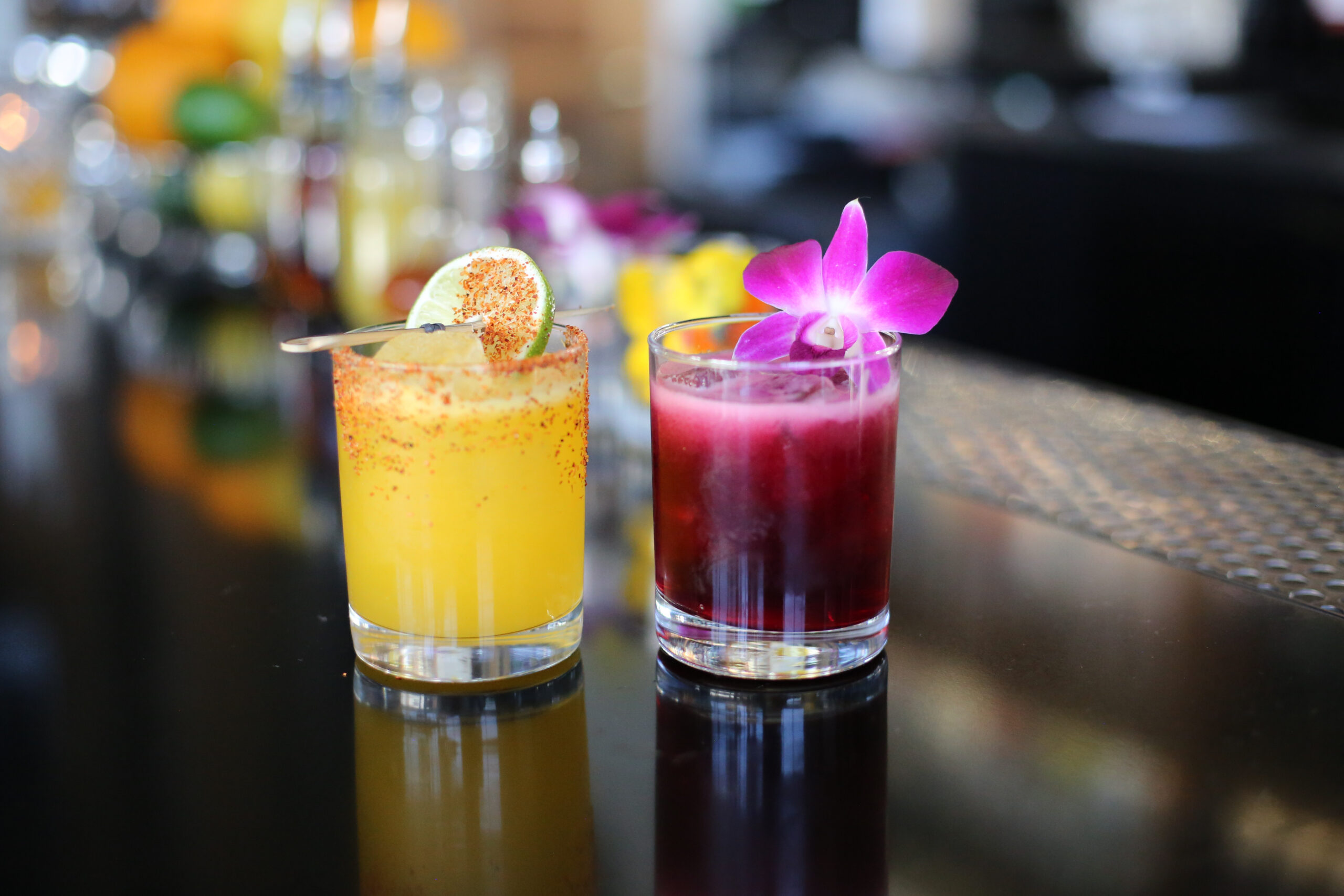 Two vibrant cocktails on a bar counter, one yellow drink garnished with a lime slice and chili powder rim, and the other a deep red drink adorned with a purple orchid.